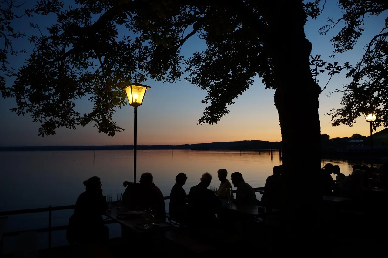 Biergarten am Abend: Gäste an langen Tischen unter Kastanien am Seeufer